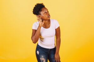 A confident African American woman poses with a listening gesture against a vibrant yellow background.